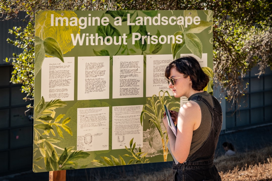 Image of a person standing to the right of a sign which says 'Imagine a Landscape Without Prisons.'