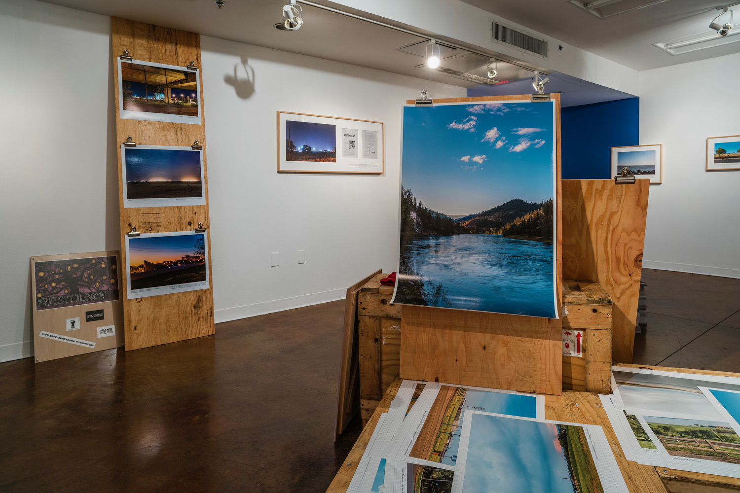 Installation of framed and unframed landscape photographs, stacks of newspaper, and shipping crates.