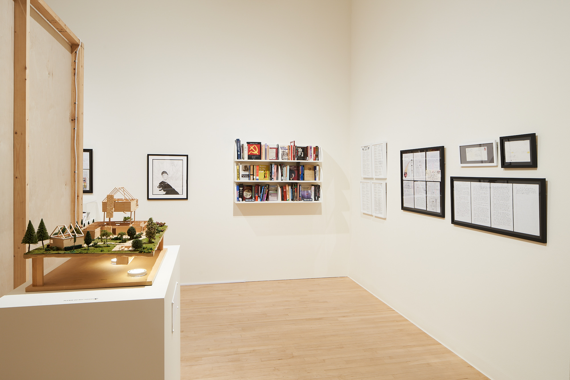 Installation image of a model house, several framed letters, and a bookshelf with books.