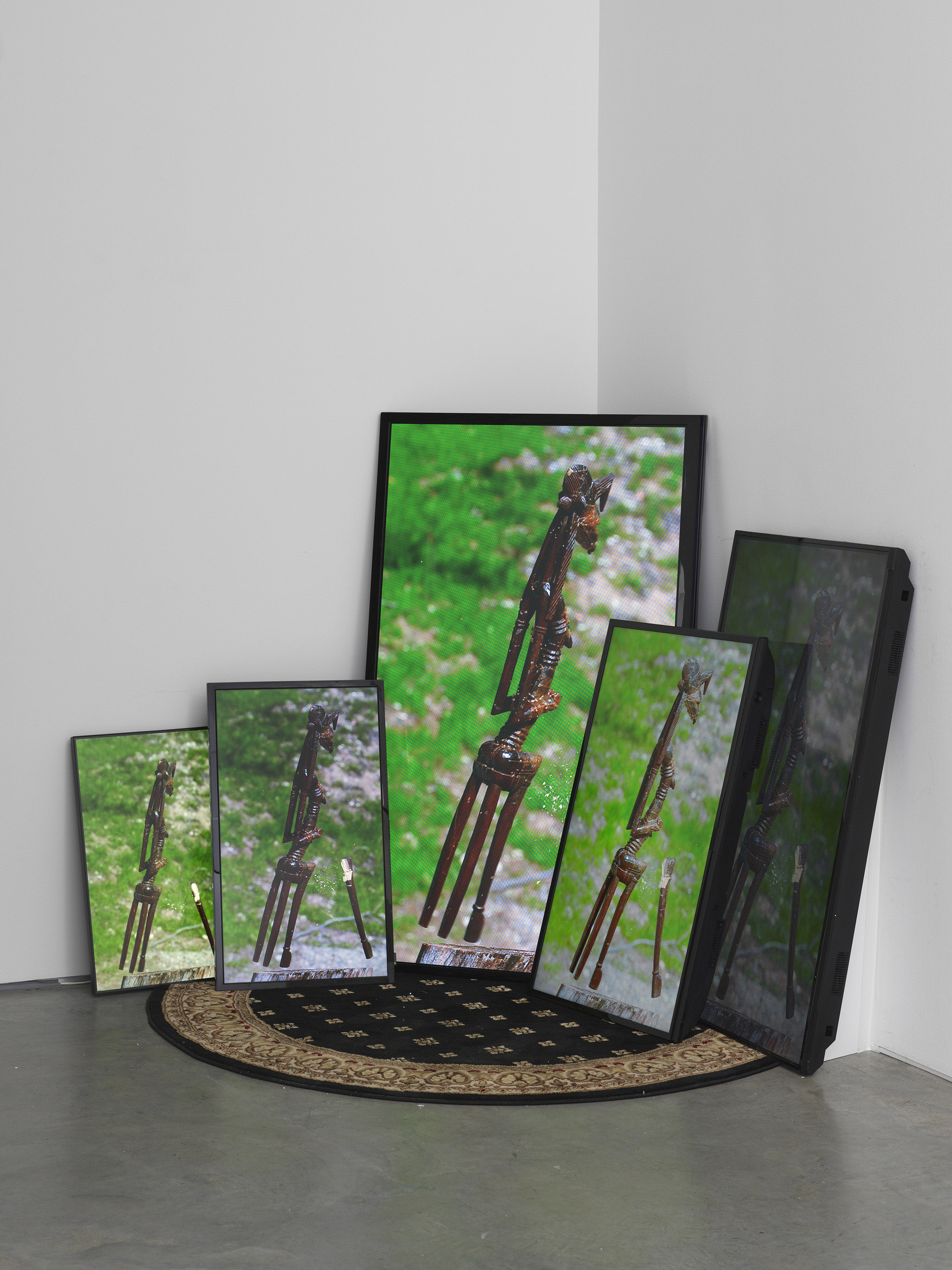  Image of five television screens leaning against a wall on a black carpet. The screens show a wooden statue from the African continent